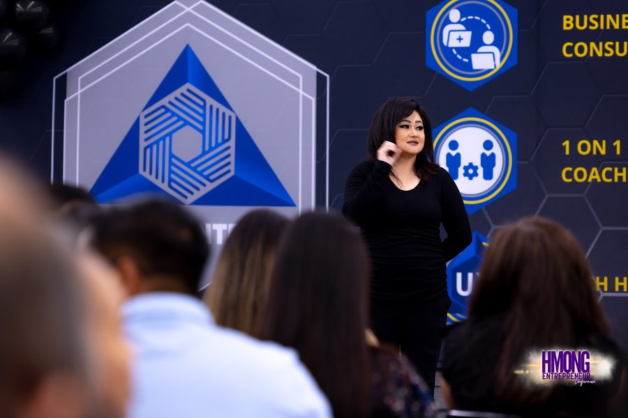 A speaker presenting to a seated audience beside a banner reading business consulting and one-on-one coaching.